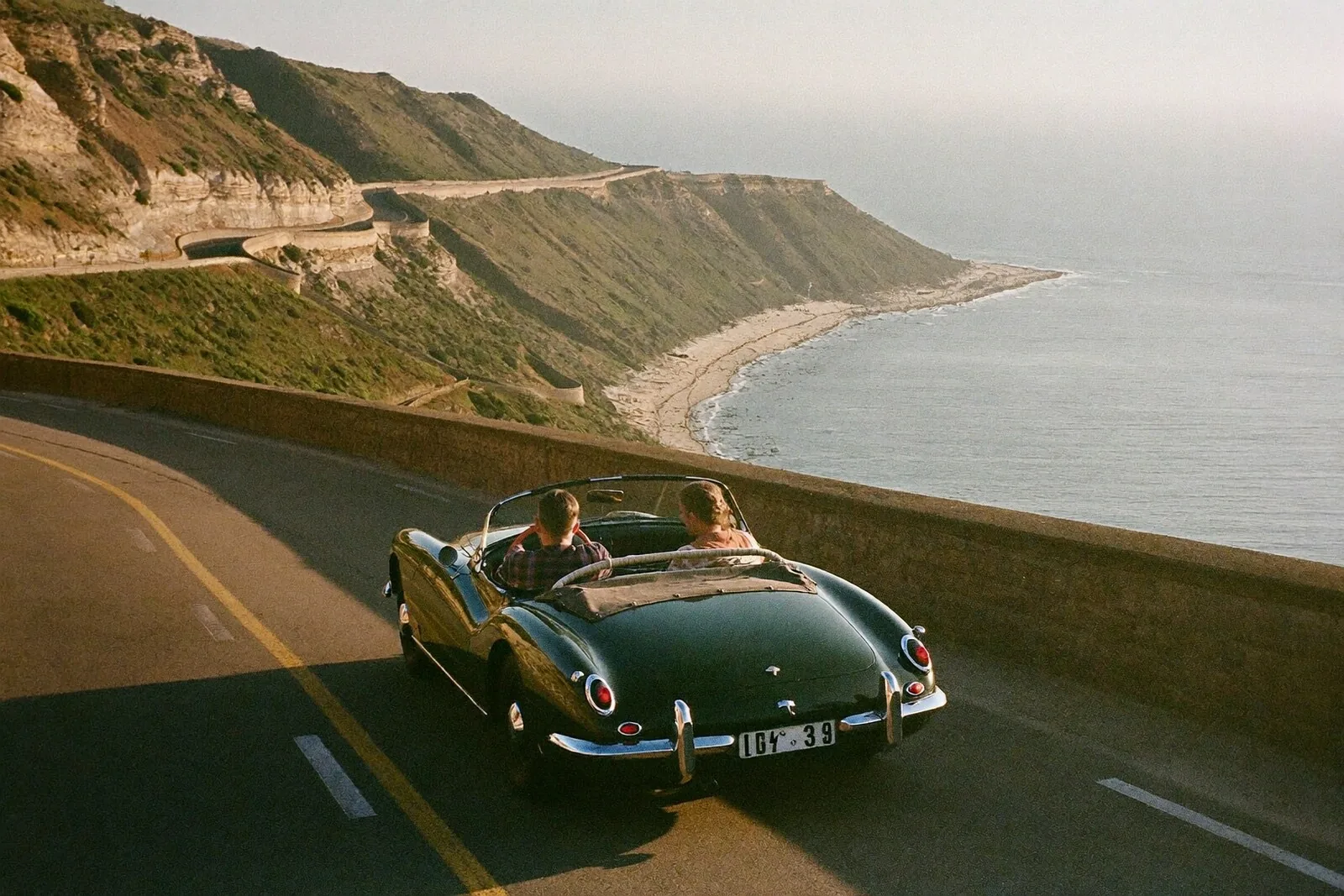 Green classic roadster driving a cliffside coastal road above the sea at golden hour