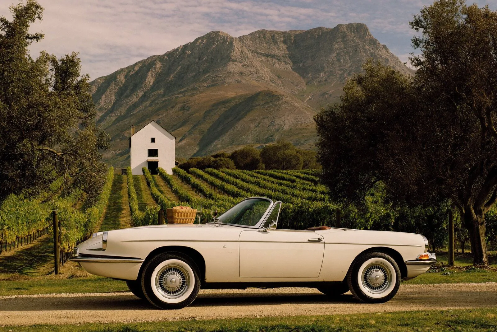 Ivory classic convertible parked beside vineyards with mountains and a Cape Dutch farmhouse behind